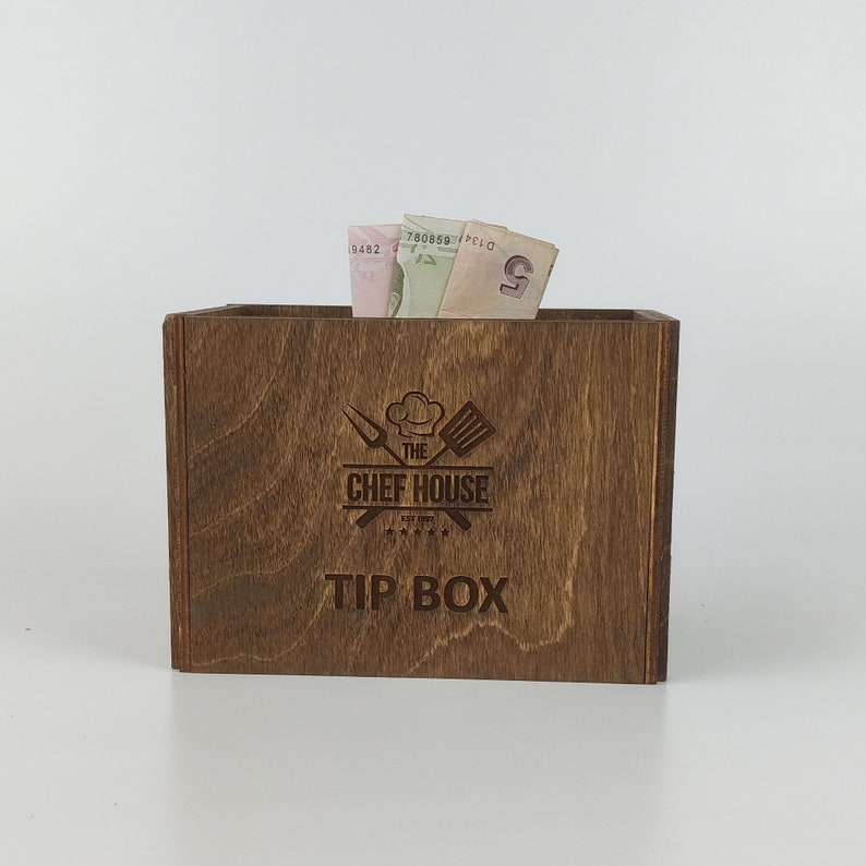 May include: A wooden tip box with the text "The Chef House" and "Tip Box" engraved on the front. The box is brown and has a rustic look.
