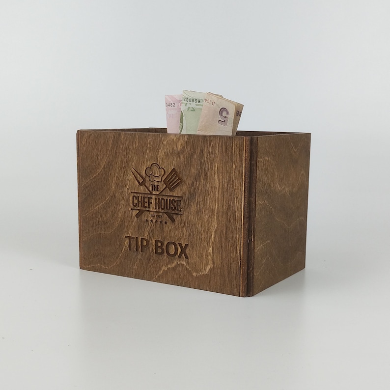 May include: A brown wooden tip box with the text "The Chef House" and "Tip Box" engraved on the front. The box has a slot for inserting money.