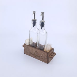 Może przedstawiać: Drewniany dozownik z dwoma szklanymi butelkami z nalewakami, przegrodą na wykałaczki i przegrodą na serwetki. Dozownik ma wygrawerowane słowo "Restaurant" z przodu.