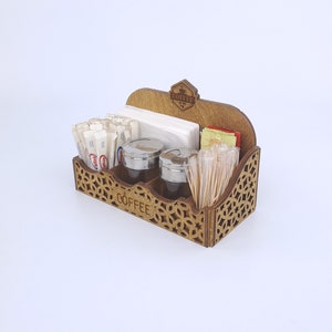 May include: Wooden coffee and condiment organizer with a decorative cutout design. The organizer has compartments for sugar packets, stir sticks, napkins, and two small containers with silver lids. The word "COFFEE" is visible on the front.