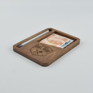 May include: A wooden bill and pen holder with a slot for a pen and a pocket for bills. The holder has a brown stain and the text "RESTAURANT GRILL HOUSE EST. 1990" is engraved on the front.