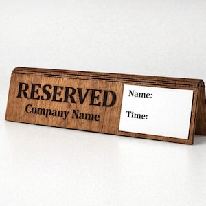 Może przedstawiać: Drewniany znak na stół z wygrawerowanym słowem „REZERWACJA” i „Company Name”. Posiada puste białe miejsce na wpisanie „Imię:” i „Czas:”. Znak jest brązowy i stoi na białej powierzchni.