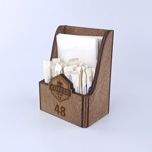 Könnte beinhalten: Holz-Theken-Zucker- und Serviettenspender mit brauner Oberfläche. Der Spender trägt ein Schild mit der Aufschrift "Coffee Est. 1997" und die Nummer "48".