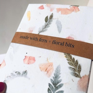 May include: A white notebook with a brown paper label that says "made with fern + floral bits". The notebook cover is made of recycled paper with pressed ferns and flower petals.