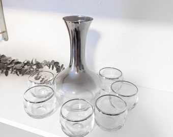 May include: A set of silver-plated glassware, including a decanter and six glasses. The decanter has a wide base and a narrow neck. The glasses are small and have a silver rim.