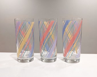 May include: Three clear glass tumblers with a colorful diagonal plaid pattern in pastel shades of pink, yellow, blue, and purple. The glasses are cylindrical and appear to be vintage or retro-style drinkware.