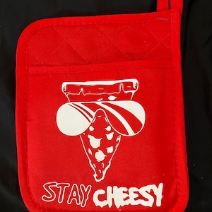 May include: Red quilted oven mitt with a white graphic of a pizza slice wearing sunglasses. The text "STAY CHEESY" is printed below the graphic. Includes a hanging loop.