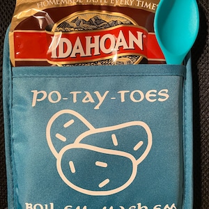 May include: A teal blue pot holder with the words "po-tay-toes" and a graphic of three potatoes printed in white. The pot holder has a pocket holding a package of Idahoan potatoes and a teal spoon. The text "BOIL EM, MASH EM, STICK EM IN A STEW" is printed on the bottom.