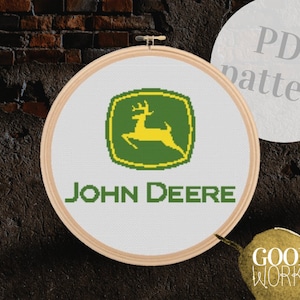 Op de afbeelding: Een kruissteekpatroon met het John Deere-logo, een groen en geel hert in een groen vierkant met de tekst "JOHN DEERE" eronder. Het patroon wordt weergegeven in een houten borduurring.