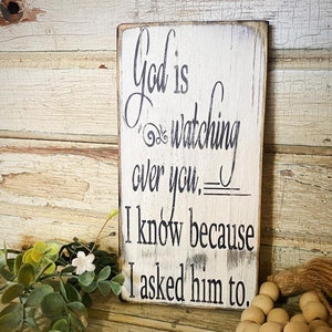May include: White wooden sign with black text that reads "God is watching over you. I know because I asked him to."