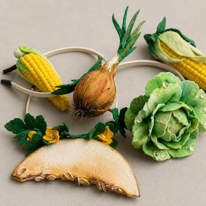 Könnte beinhalten: Eine Sammlung von Haaraccessoires mit gefilzten Gemüse- und Obstmotiven, darunter zwei gelbe Maiskolben, eine braune Zwiebel, ein grüner Kohlkopf mit klaren Tautropfen sowie eine Melonenscheibe mit kleinen gelben Blüten und grünen Blättern.