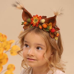 May include: A brown felt headband with attached fox ears, adorned with felt flowers, acorns, and berries in shades of orange, green, and red. The ears have fluffy, light-colored accents. The headband is worn by a young person with blonde hair.
