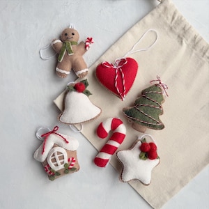 Könnte beinhalten: Eine Sammlung von sechs Filzanhängern, bestehend aus einem Lebkuchenmännchen mit grünem Schal, einem roten Herz, einer weißen Glocke mit roter Beere, einem grünen Weihnachtsbaum, einer rot-weißen Zuckerstange und einem weißen Stern mit roten Beeren.