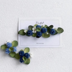 May include: Two felt hair accessories featuring clusters of dark blue berries, small white flowers, and green leaves. The larger piece is attached to a white display card with the text Pastel and Felt accessories printed in black ink.