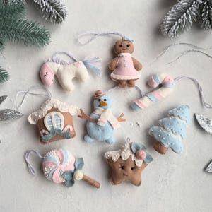 Può includere: Un set di sette decorazioni natalizie in feltro che raffigurano un omino di pan di zenzero, un pupazzo di neve, un unicorno, un bastoncino di zucchero, una casetta di pan di zenzero, un lecca-lecca e una stella, nei toni pastello rosa, blu e crema.