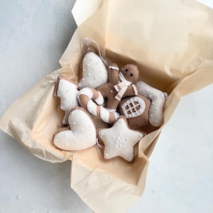 Puede incluir: Una colección de adornos navideños de fieltro hechos a mano en una caja forrada con papel kraft. Los adornos incluyen un hombre de jengibre, una estrella, un corazón, un bastón de caramelo, una casa y un ángel, todos con detalles de cuentas blancas y una combinación de colores marrón y blanco.