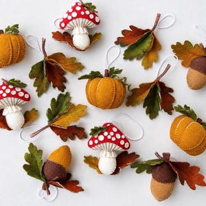 Könnte beinhalten: Eine Sammlung von Filzornamenten mit herbstlichen Motiven, darunter rot-weiß gepunktete Pilze, orangefarbene Kürbisse, braune Eicheln und mehrfarbige Eichenblätter in Grün, Gelb und Braun, jeweils mit einer weißen Schlaufe zum Aufhängen.