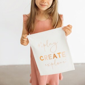 May include: A young girl in a pink dress holds a white canvas tote bag with the words "play create explore" printed in orange and pink.