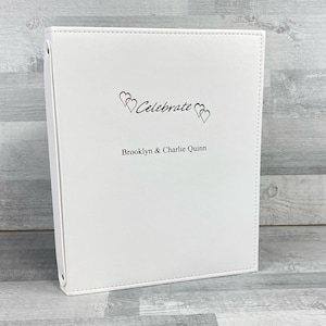 May include: White leather photo album with the text "Celebrate" and two hearts above the names "Brooklyn & Charlie Quinn".