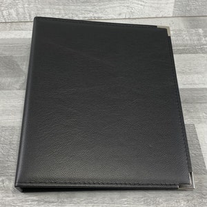 May include: A black, textured, rectangular binder featuring a smooth, leather-like finish. The front cover is accented with a single, polished silver-toned metal corner protector at the top right and another at the bottom right edge of the cover.