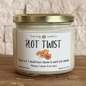 May include: A glass jar candle with a gold lid. The label reads "PLOT TWIST" with a cartoon dog and cat illustration. Text below says "People suck. I should have chosen to work with animals." The candle is 3.5 oz / 200 g.