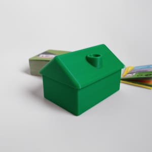 May include: A bright green house-shaped container with a chimney detail, likely for game pieces. A stack of green-backed cards and a small pile of colorful cards are visible in the background, suggesting a board game theme.