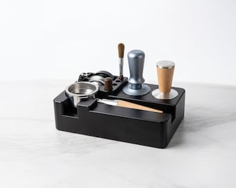 Könnte beinhalten: Eine schwarze Espresso-Station mit verschiedenen Werkzeugen zur Kaffeezubereitung. Sie enthält einen Siebträger, einen Tamper, ein Verteilwerkzeug, eine Bürste und einen Tamper mit Metall- und Holzgriff. Die Station dient dazu, den Kaffeezubereitungsprozess zu organisieren und zu rationalisieren.