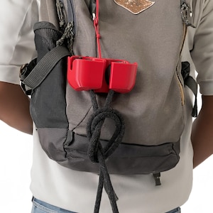 Könnte beinhalten: Ein rotes Plastiketui mit einem schwarzen Band, das an einem grauen Rucksack befestigt ist. Das Etui hat zwei Fächer und hängt oben am Rucksack.