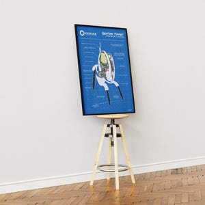 May include: A framed blueprint-style poster featuring a diagram of a "Sentient Turret" from the video game Portal. The poster is in a black frame and is displayed on a wooden and metal stool against a white wall.