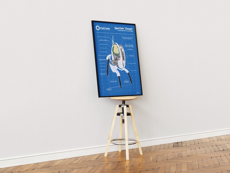 May include: A framed blueprint-style poster featuring a diagram of a "Sentient Turret" from the video game Portal. The poster is in a black frame and is displayed on a wooden and metal stool against a white wall.