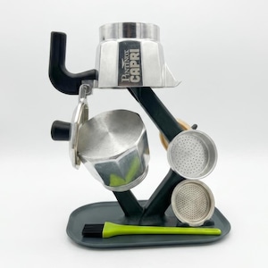 Puede incluir: Una cafetera espresso plateada y negra con el texto "Pintinox Capri" en el lateral. La cafetera espresso está sobre un soporte verde oscuro con un cepillo verde y dos filtros plateados.