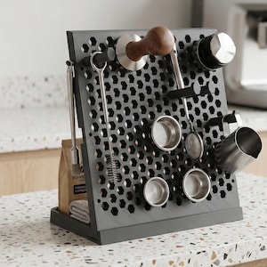 Op de afbeelding: Een grijze koffiestation-organizer met een honingraatpatroon, met diverse roestvrijstalen koffiegereedschappen, waaronder een stamper, een portafilter en een melkopschuimer. Een zak koffiebonen en een kleine handdoek zijn ook zichtbaar.