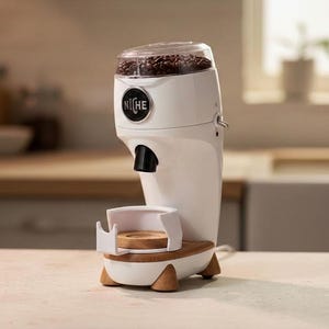 以下が含まれることがあります： コーヒー豆が入った透明なトップが付いた白い電動コーヒーグラインダー。グラインダーは木製のベースと黒い注ぎ口が付いています。「Niche」のブランド名が前面に表示されています。グラインダーは明るい色のカウンタートップにあります。