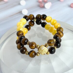 May include: Two beaded bracelets. One bracelet features brown and gold tiger eye beads, the other has yellow beads. The bracelets are connected by a gold-coloured decorative piece with a pearl in the centre. The bracelets are displayed on a clear, round stand.