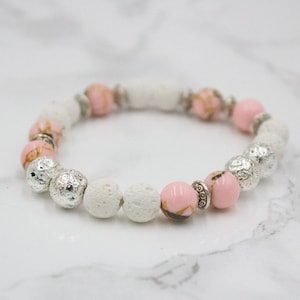 May include: A bracelet with a mix of white, pink, and silver beads. The bracelet has textured silver beads, smooth pink beads, and white lava rock beads. The design is strung on a stretchy cord, creating a stylish accessory.