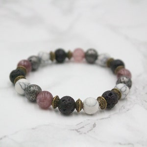 May include: A beaded bracelet with black lava rock, white and gray stones, and pink quartz beads. The beads are separated by small gold-toned spacers.
