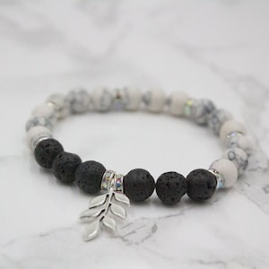 May include: A beaded bracelet featuring a mix of white and gray patterned beads, interspersed with black lava beads. A silver-colored leaf charm hangs from the bracelet. The bracelet is designed for everyday wear.