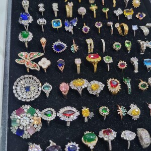 Puede incluir: Una colección de anillos variados con piedras preciosas coloridas y diseños intrincados. Los anillos presentan una variedad de formas, incluyendo piedras redondas, cuadradas y en forma de pera, engastadas en bandas de oro y plata. Las piedras preciosas incluyen tonos azules, verdes, amarillos y rojos.