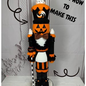 May include: A Halloween-themed nutcracker with a pumpkin face, black and orange striped trousers, and a black hat topped with a white and orange candy-themed decoration. The nutcracker is holding a candy cane-style staff. The text "LEARN HOW TO MAKE THIS" is written in white on a black background.