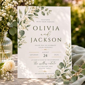 Puede incluir: Una tarjeta de invitación de boda rectangular con hojas de eucalipto en acuarela verde oscuro y pequeñas flores blancas. El texto dice: Olivia and Jackson, Saturday August 24 2026, at four o'clock in the afternoon, The Willow Estate, 2675 Willow Lane, Austin, Texas.
