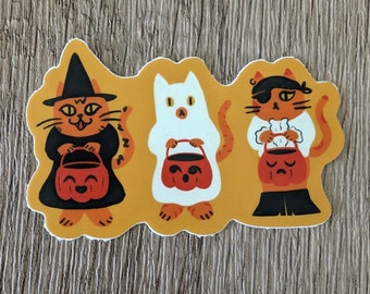 May include: Three orange cats dressed up for Halloween. One cat is wearing a witch's hat, another is a ghost, and the third is a pirate. Each cat is holding a pumpkin-shaped trick-or-treat bag.