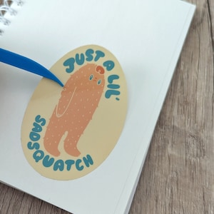 May include: A light brown oval sticker featuring a cartoon illustration of a furry creature with a sad expression. The text "Just a lil' Sasquatch" is written in a blue and yellow font around the creature.