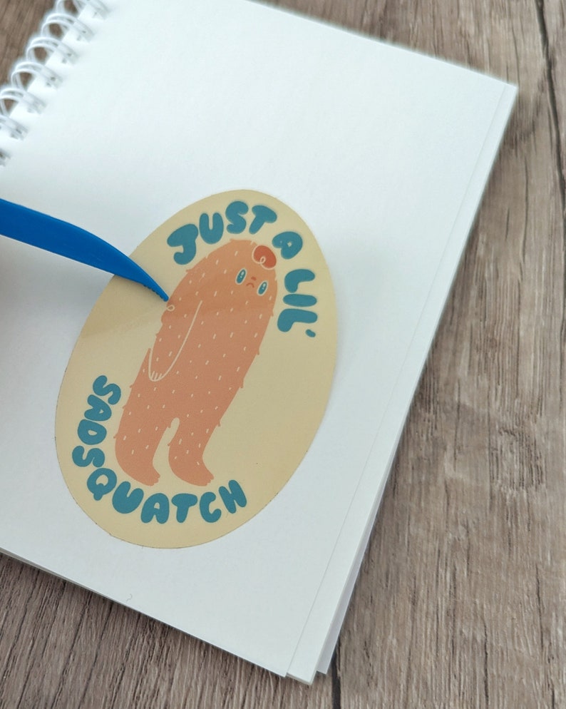 May include: A light brown oval sticker featuring a cartoon illustration of a furry creature with a sad expression. The text "Just a lil' Sasquatch" is written in a blue and yellow font around the creature.