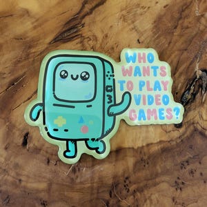 May include: A light green cartoon character pin with a smiling face and the text "WHO WANTS TO PLAY VIDEO GAMES?" in pink, blue, and yellow. The pin is on a wooden surface.
