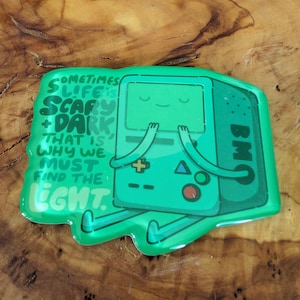 May include: A green BMO-shaped pin with the text "Sometimes life is scary + dark that is why we must find the light." The pin features a smiling face and arms. The pin is on a wooden surface.