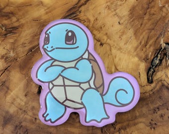 May include: A light blue Squirtle character pin with a brown shell and a lavender border. The pin features the Squirtle character with its arms crossed. The pin is on a wooden surface.