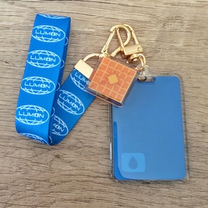 May include: A blue lanyard with the word "LUMON" repeated multiple times. Attached is a gold-colored clasp holding a small waffle-patterned charm and a blue ID holder with a water droplet icon.