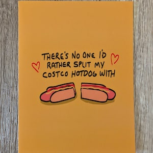 May include: An orange greeting card with the text "There's no one I'd rather split my Costco hotdog with" in black lettering. Two cartoon hotdogs are below the text, with red hearts on either side.