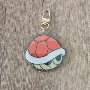 May include: A red and white turtle shell keychain with a gold-colored clasp. The shell has a segmented design with a white border and a cartoonish eye detail. The keychain is designed to be attached to keys or a bag.