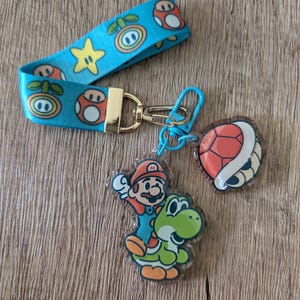 May include: A blue and green keychain with a Mario and Yoshi character charm, a red and brown turtle shell charm, and a blue and gold clip. The keychain has a blue strap with a repeating pattern of mushrooms, stars, and plants.